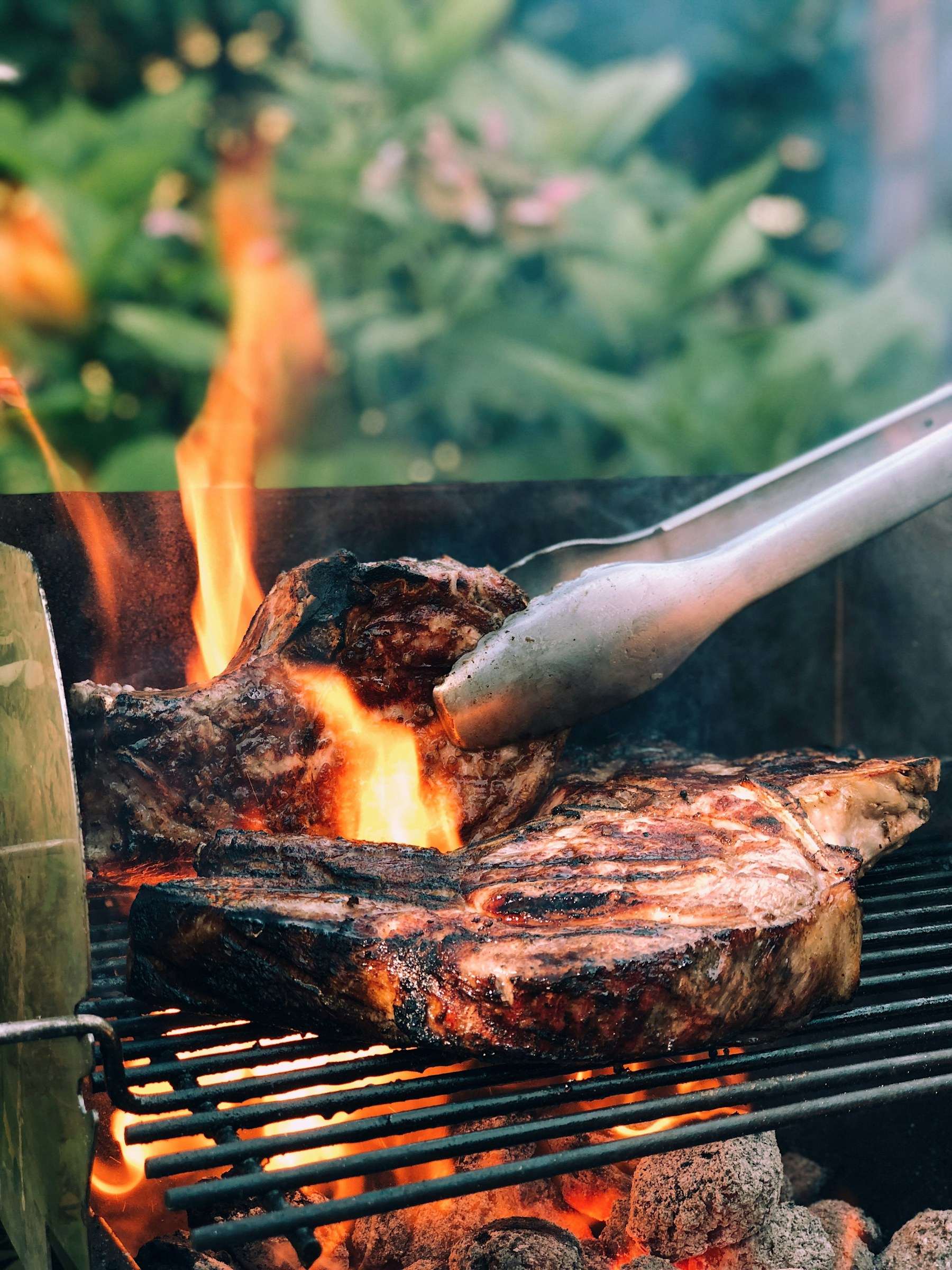 Unas pinzas levantan un filete sobre la parrilla, con la llama de la brasa debajo